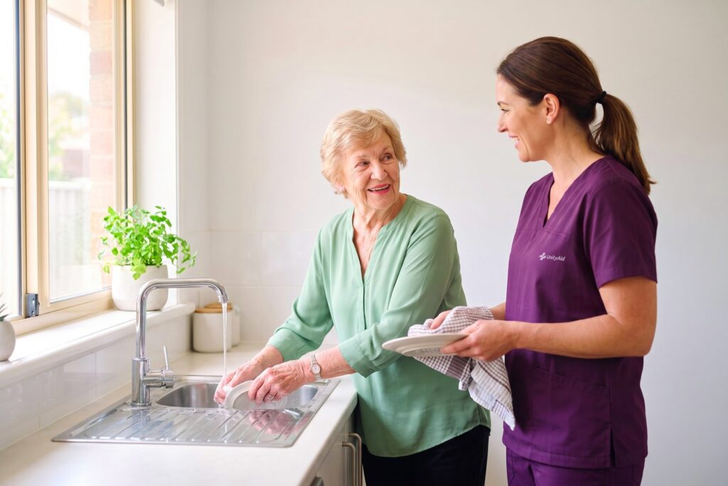 An older woman washing up at her sink while a support worker dries beside her.